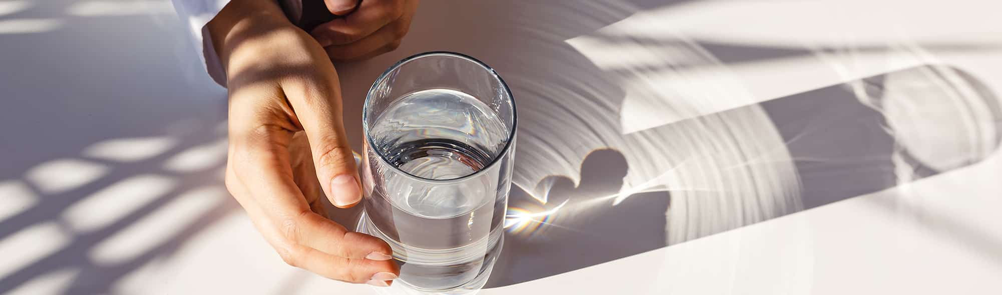 Woman's hands and a glass of clean water on the white table in natural sunlight with plant shadows.